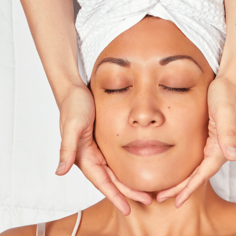 Esthetician administering a facial massage to a client, who is wearing a towel on her hair and looks relaxed